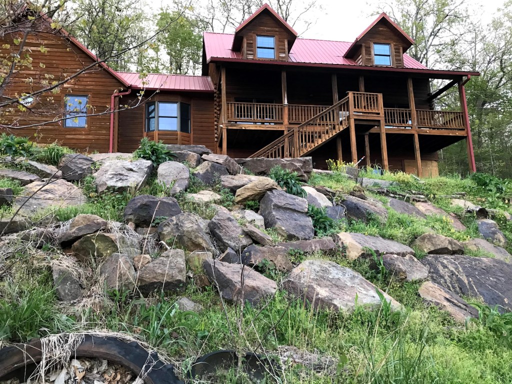 The log house seen from below, with a wide cascade of mostly bare boulders and only a few patches of green on the slope in front