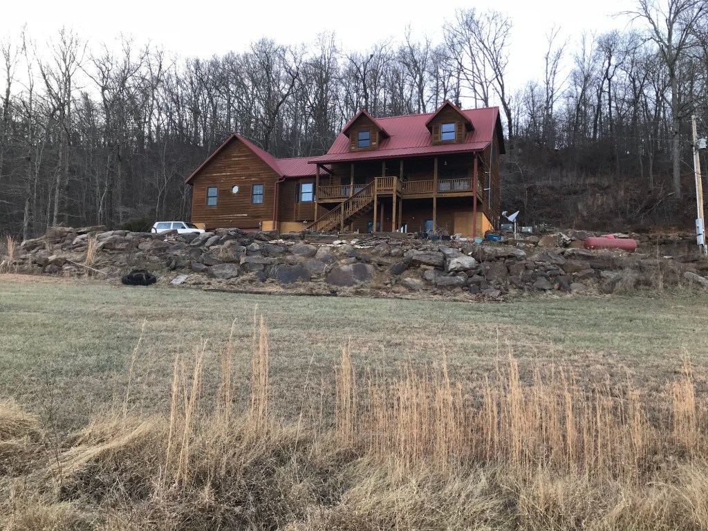 The log house in winter seen from below, with a bare boulder wall and plain lawn in front and no gardens