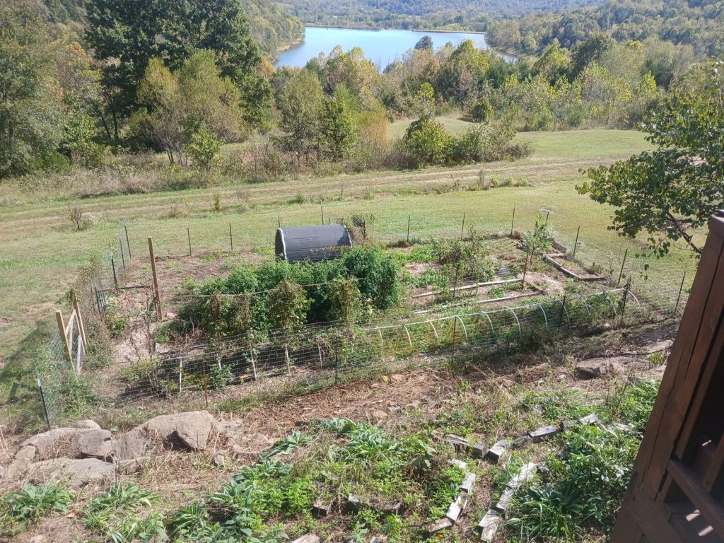 A fenced vegetable garden with white low-tunnel hoops below the log house deck, with comfrey in front and Prayer Lake and wooded hills in the distance
