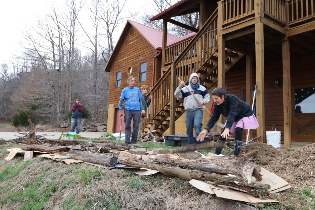 A group of people laying logs over cardboard on the slope in front of the log house to start a garden bed