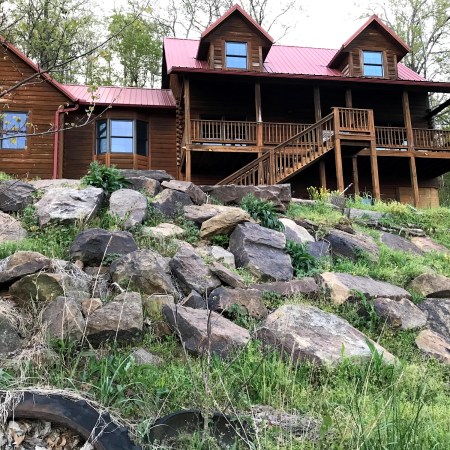 The cypress log house with its red metal roof, two dormers, and covered front porch, seen from below across a rocky, grassy slope