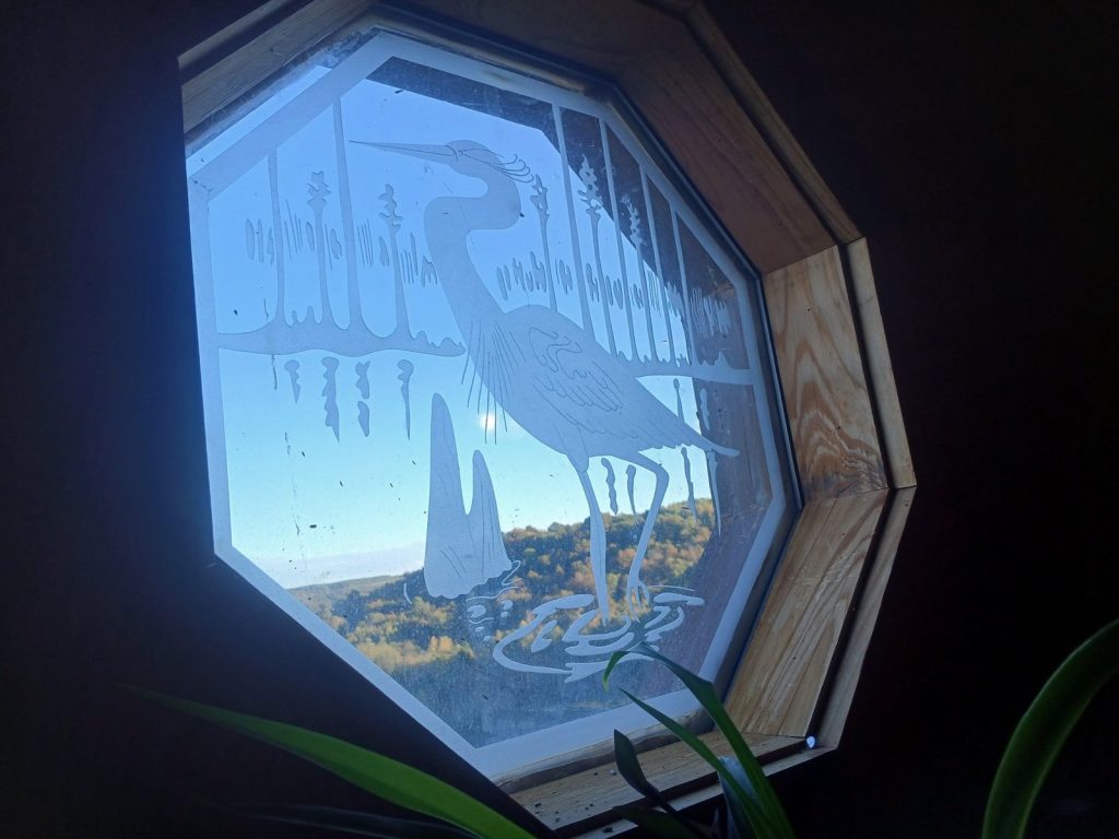 Octagon window with an egret etched into the glass, with wooded Ozark hills in fall color and blue sky behind it