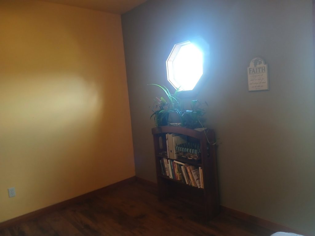 Small bookshelf with plants on top under a glowing octagon window, with a Faith sign on the wall