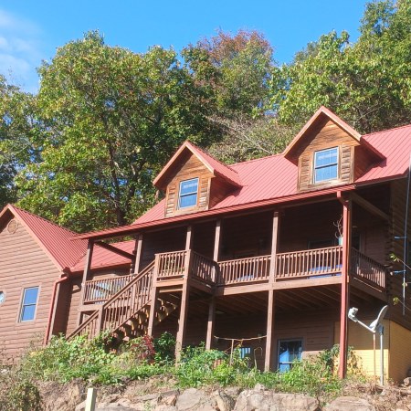 The Living Springs Log House, a log home with a red metal roof, two dormers, and a long wraparound deck, sitting above a green garden slope under tall oaks and a blue sky