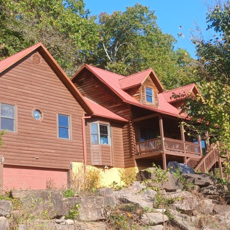 The Living Springs Log House seen from below a ledge of large Ozark boulders, with the bay window, garage, and deck under a red metal roof