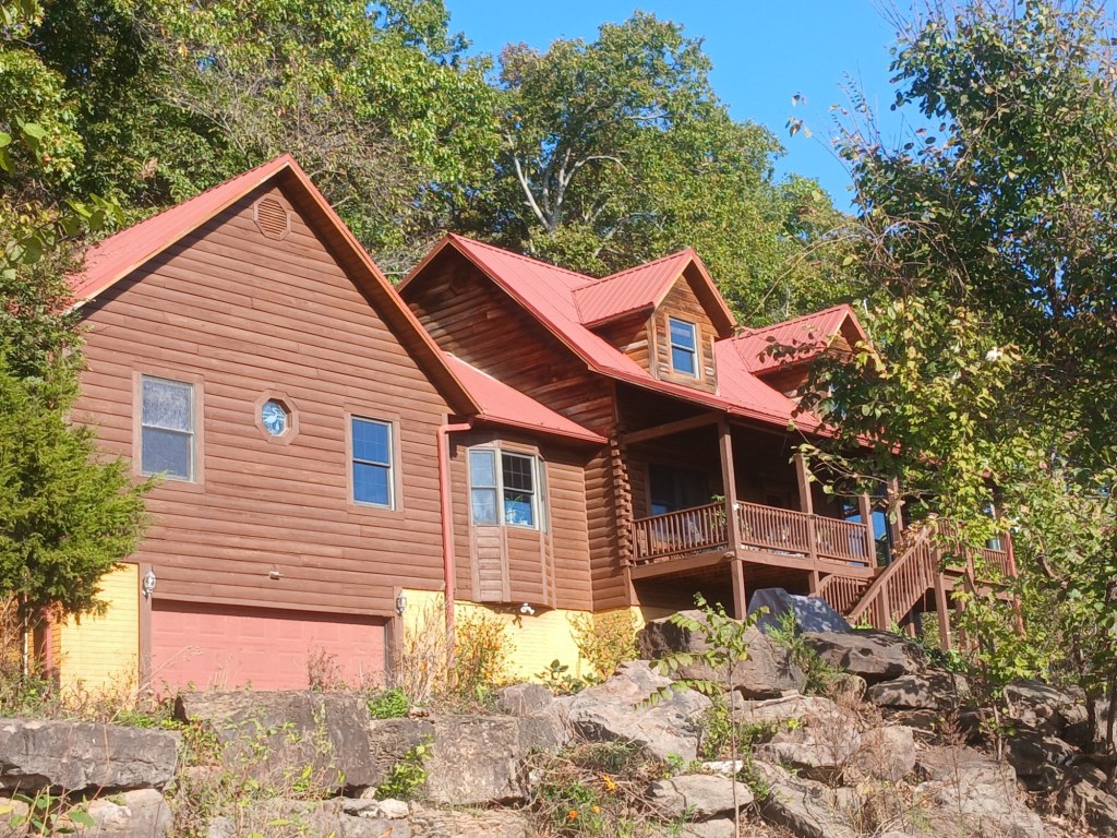 The Living Springs Log House seen from below a ledge of large Ozark boulders, with the bay window, garage, and deck under a red metal roof
