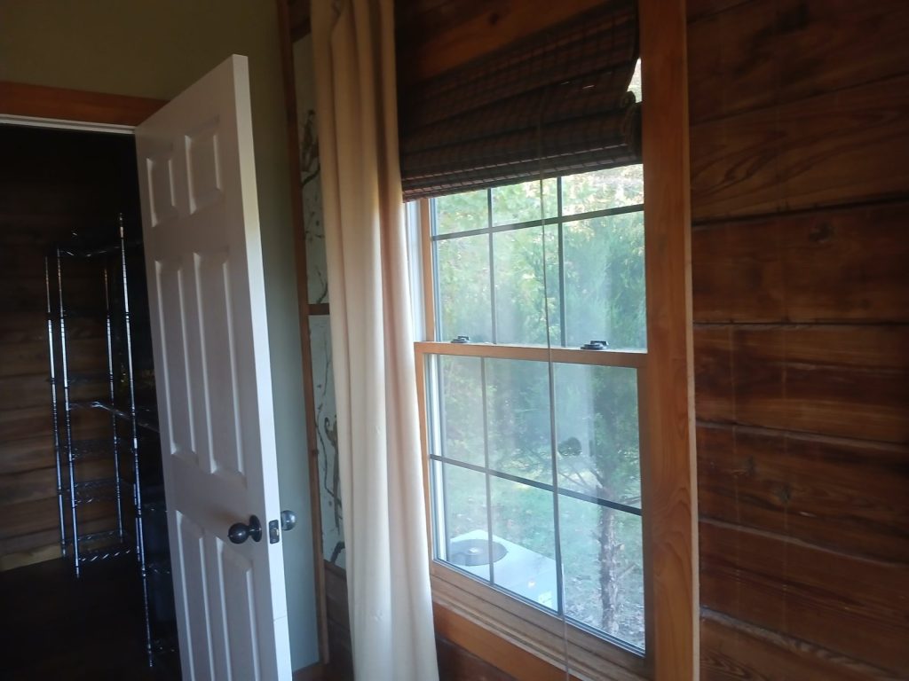 Bedroom window with the bamboo shade rolled up to green trees outside, next to an open closet with wire shelves