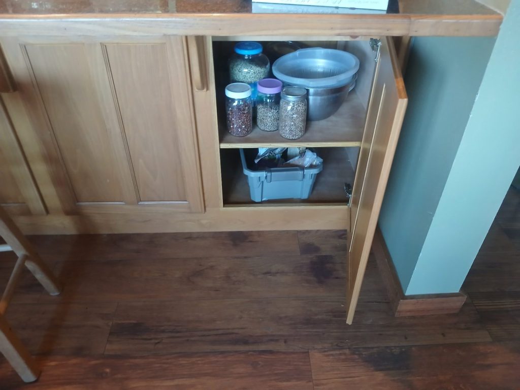Open cabinet under the kitchen bar holding jars of beans and grains, bowls and a storage bin