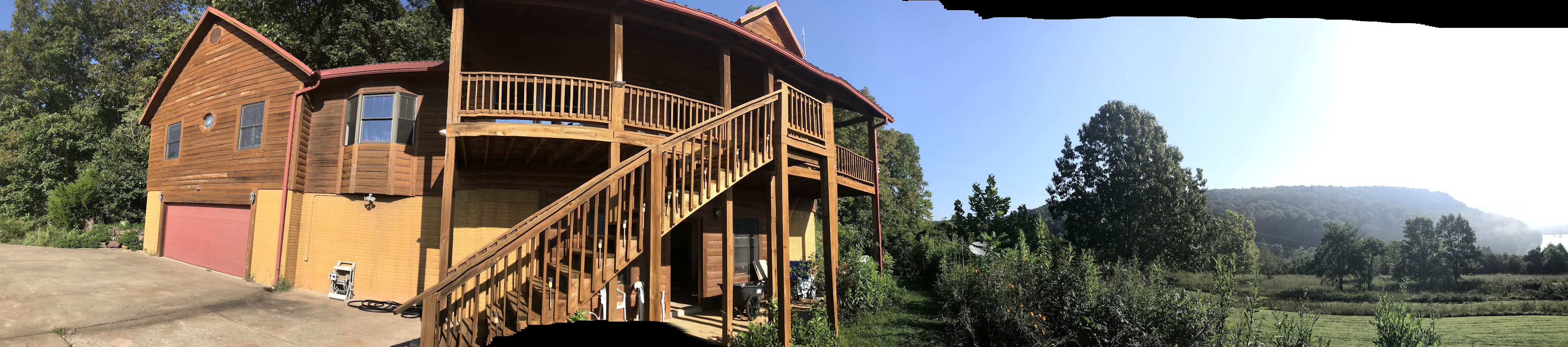 A panoramic view of the cypress log house with its long front stairs and porch, opening out to green meadows, trees, and misty Ozark hills under a blue sky