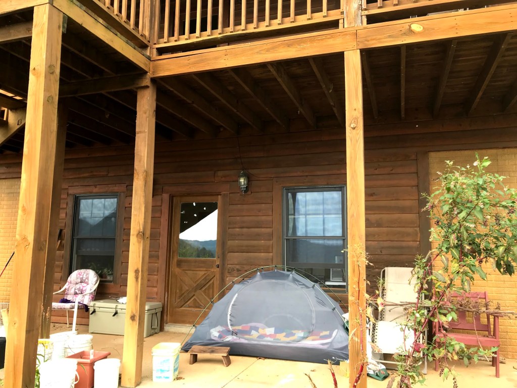 A mesh tent set up on the covered patio outside the basement suite, under the deck above, with chairs and plants nearby